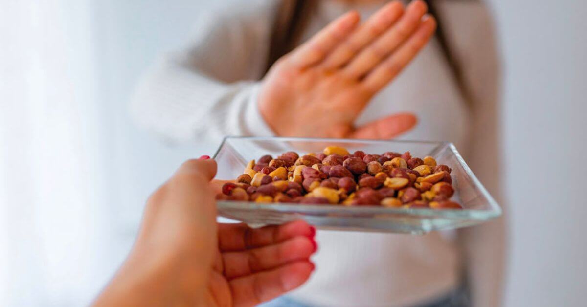 a woman with a food allergy is refusing a dish of almonds that another woman's hand is offering her